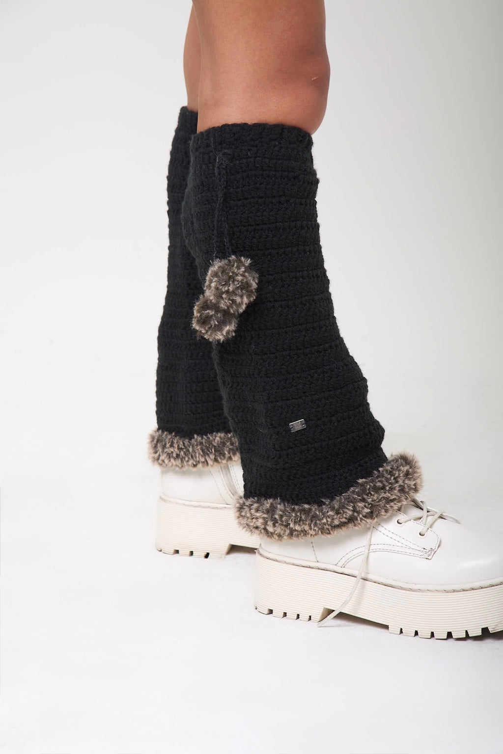 Side view of black knitted Not Your Baby legwarmers with fur trim worn over white sneakers
